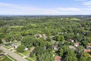Aerial view of a suburban Minnesota neighborhood with many houses surrounded by dense green trees and forests, under a bright blue sky with scattered clouds—a perfect scene for your next home for sale with Beekeeper Realty.