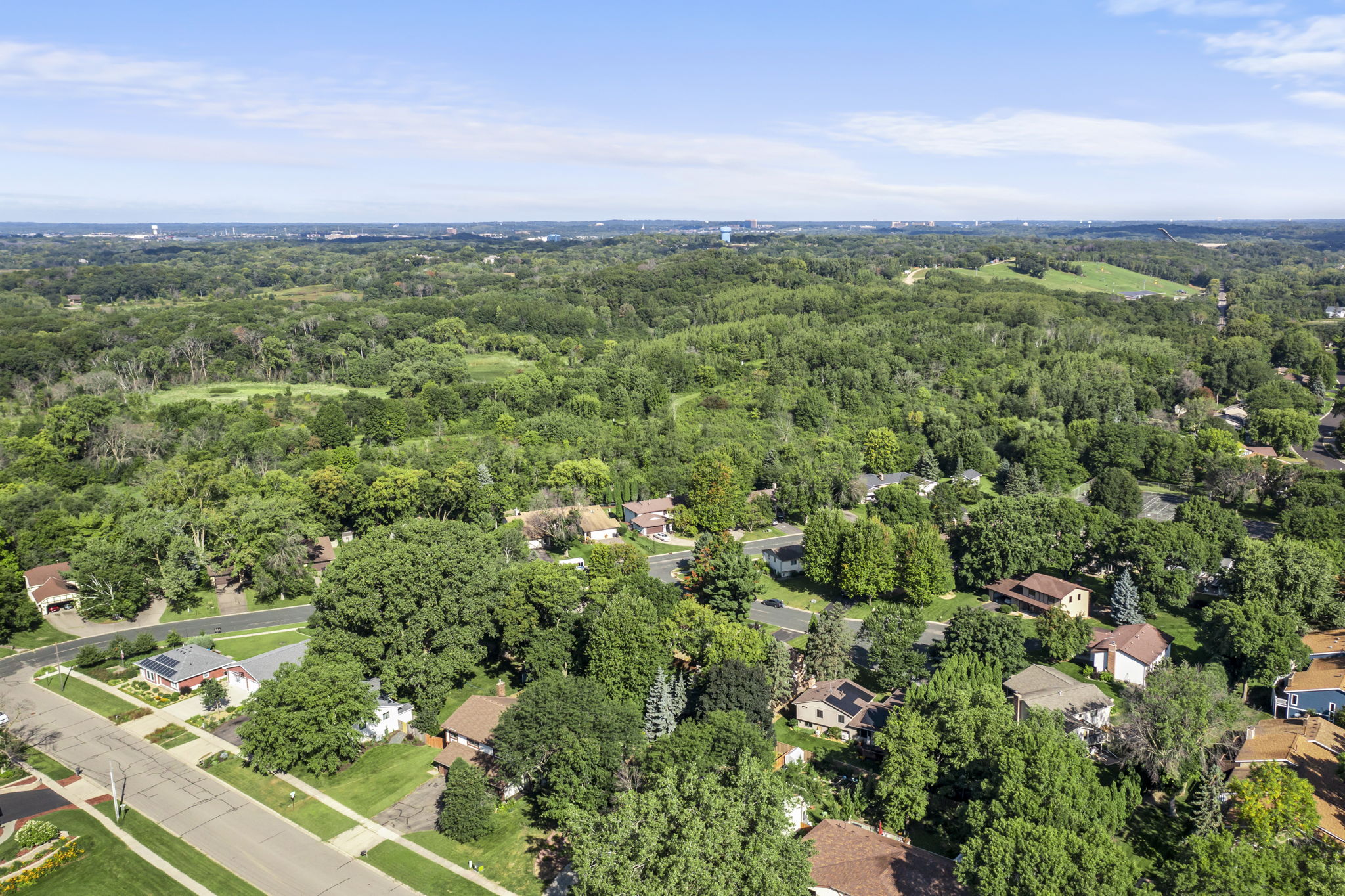 Aerial view of a suburban Minnesota neighborhood with many houses surrounded by dense green trees and forests, under a bright blue sky with scattered clouds—a perfect scene for your next home for sale with Beekeeper Realty.