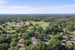 Aerial view of a Bloomington, Minnesota neighborhood with houses, tree-lined streets, a school building, parking lot, and a large green sports field with a baseball diamond—showcased by Beekeeper Realty under a clear blue sky.