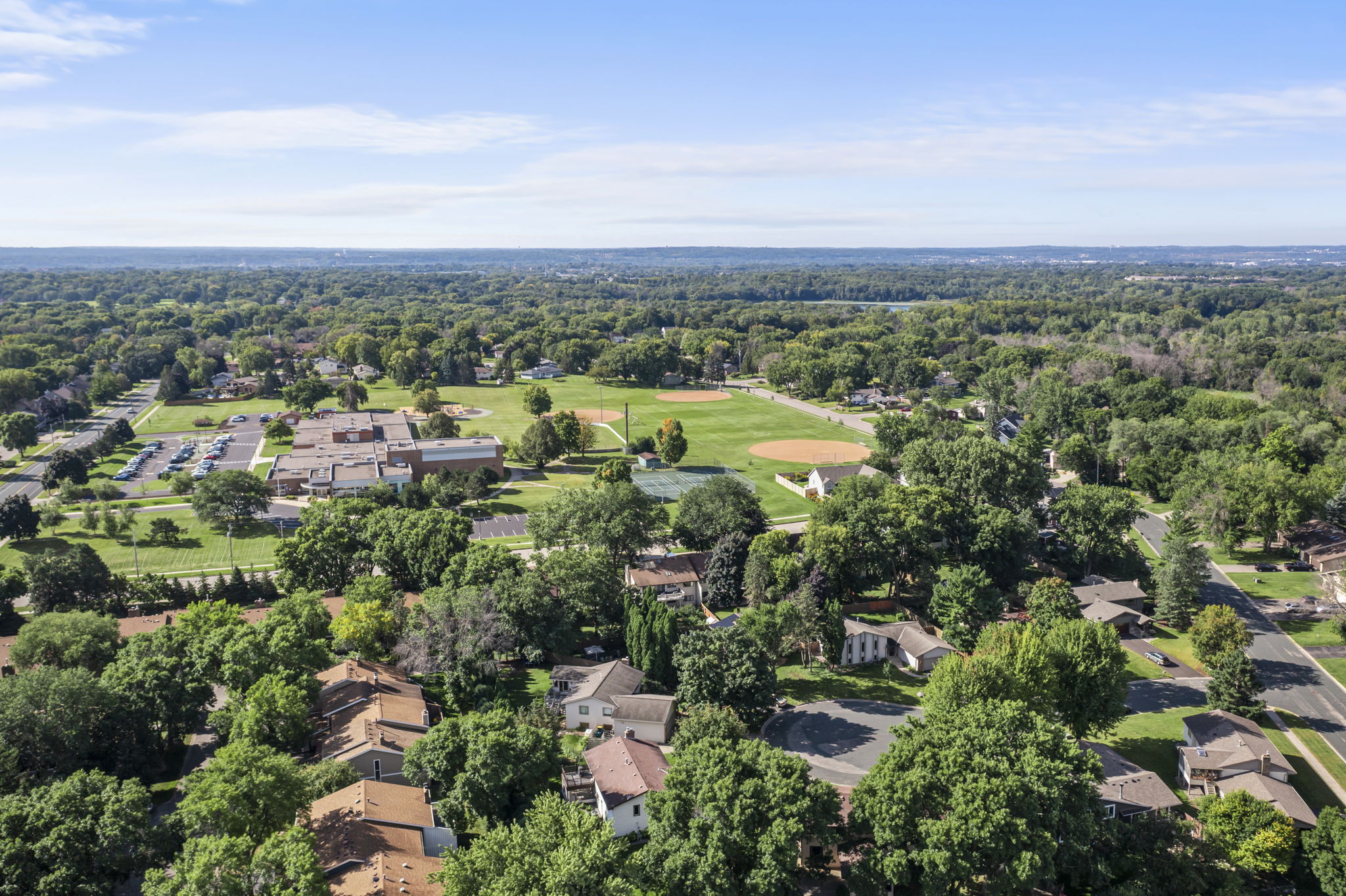 Aerial view of a Bloomington, Minnesota neighborhood with houses, tree-lined streets, a school building, parking lot, and a large green sports field with a baseball diamond—showcased by Beekeeper Realty under a clear blue sky.
