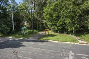 A paved path leads into a wooded area near 9333 Colorado Circle, Bloomington, with a yellow two-way arrow sign at its entrance. Grass, trees, street signs, and Beekeeper Realty signage are visible along the slightly worn road.