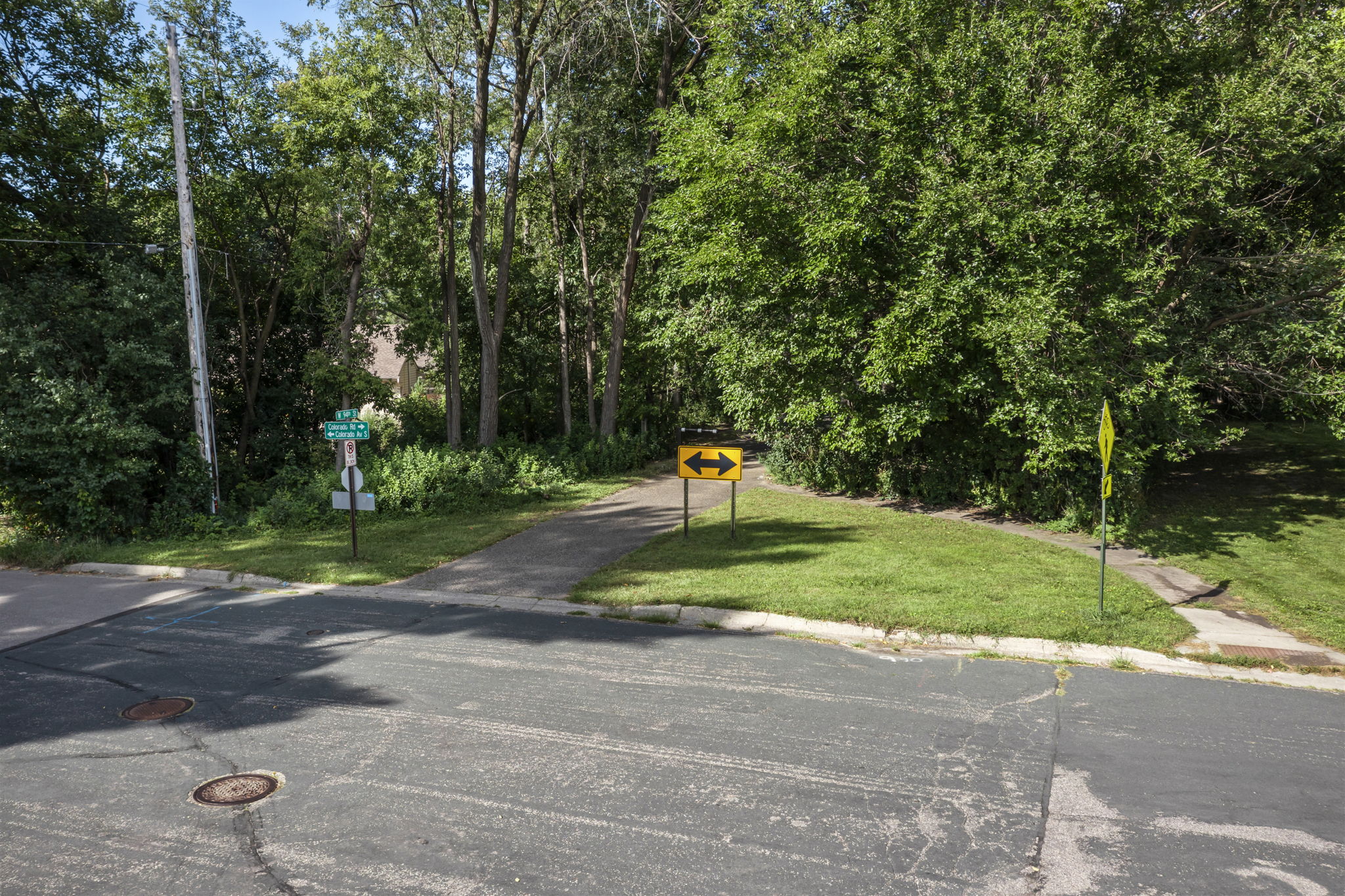 A paved path leads into a wooded area near 9333 Colorado Circle, Bloomington, with a yellow two-way arrow sign at its entrance. Grass, trees, street signs, and Beekeeper Realty signage are visible along the slightly worn road.