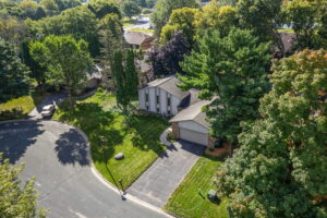 Aerial view of a Bloomington, Minnesota home for sale with a two-car garage, lush trees, green lawn, and neighboring homes along a quiet cul-de-sac with nearby parked cars.