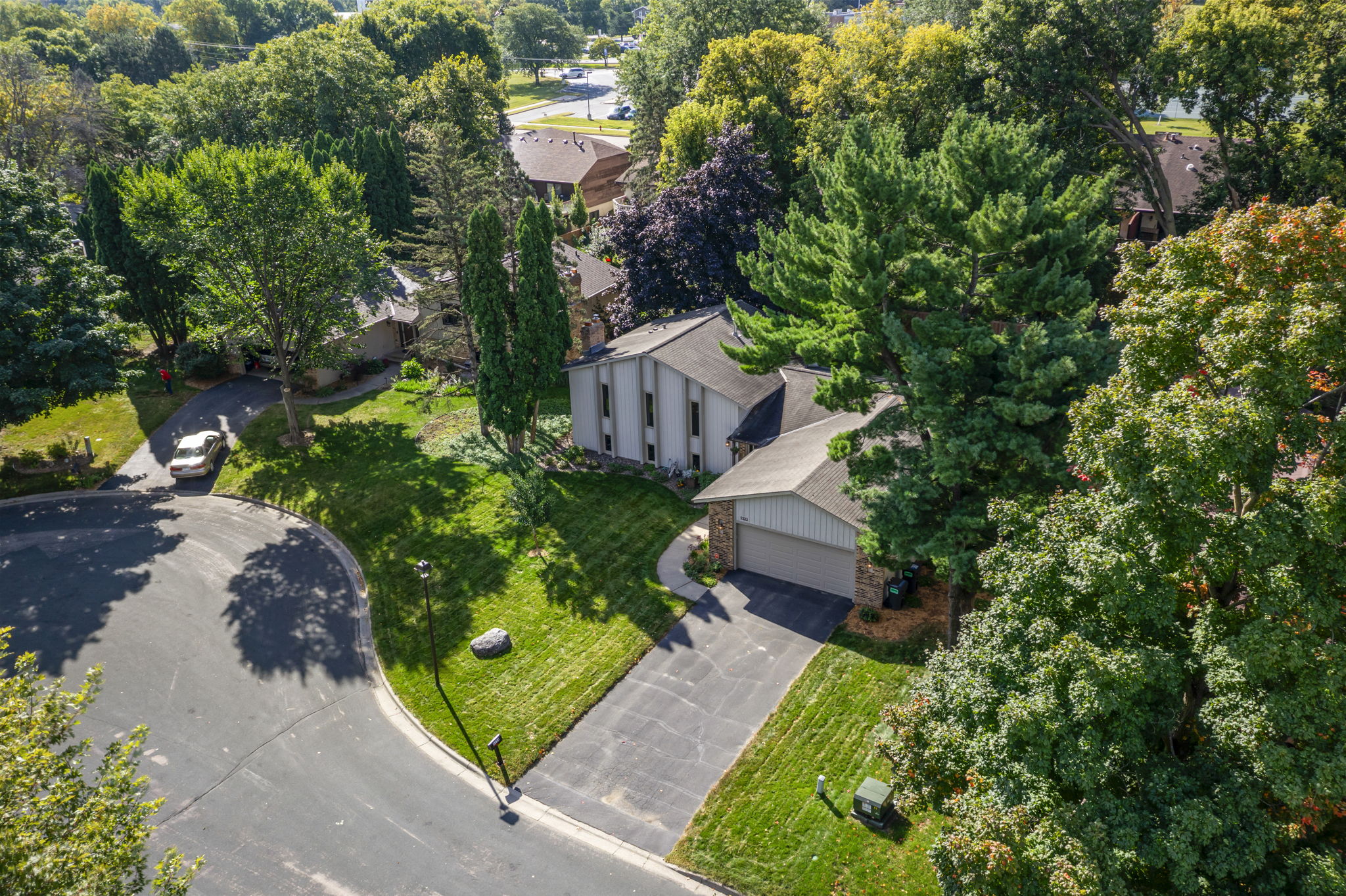 Aerial view of a Bloomington, Minnesota home for sale with a two-car garage, lush trees, green lawn, and neighboring homes along a quiet cul-de-sac with nearby parked cars.
