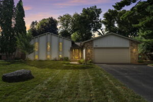 A modern two-story home for sale at 9333 Colorado Circle, with a blend of vertical siding and brick exterior, is warmly lit among trees at dusk. Manicured lawn, large rock, and a driveway to the double garage complete this Beekeeper Realty listing.