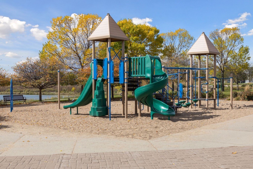 A playground with green slides and climbing equipment stands on sand. Trees with autumn leaves and a lake are in the background. The sky is partly cloudy, and there are benches near the playground.