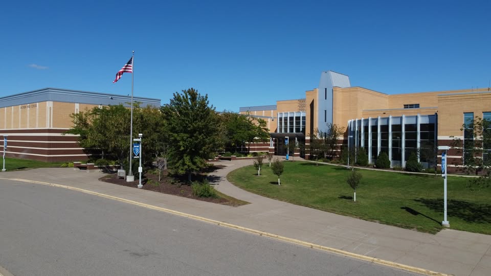 A modern school building with tan brick and large windows sits at 1648 Whitehall Place, Shakopee, MN, surrounded by green lawns and trees, with a U.S. flag flying on a pole beneath a clear blue sky.