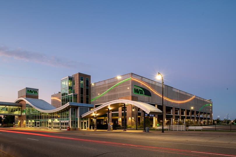 A modern Apple Valley MN transit center building features green MTA logos, curved white awnings, a glass entrance, and lights outlining the structure, photographed at dusk with a clear sky and light traffic in front.