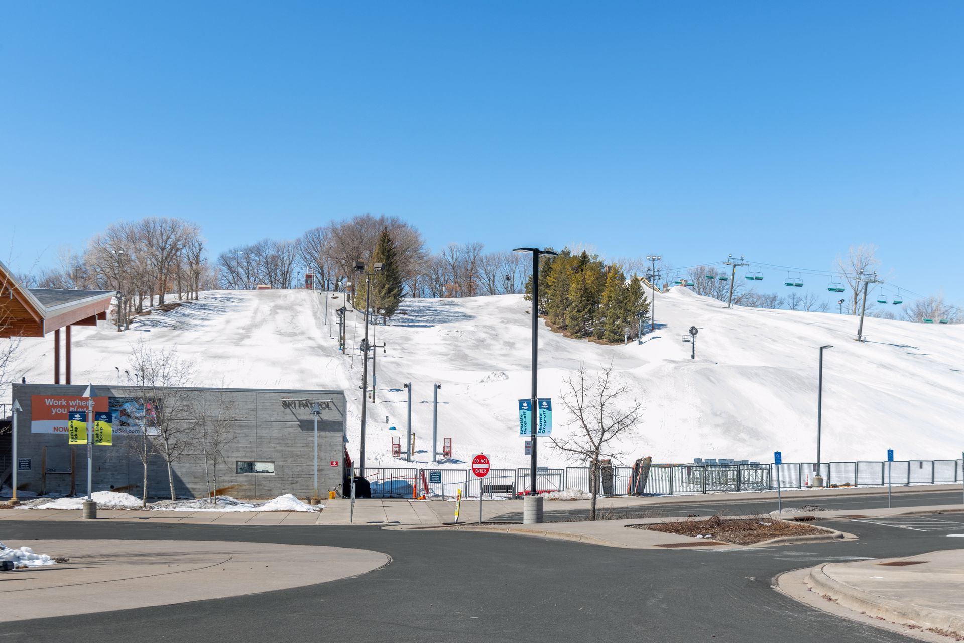 A snow-covered ski slope with a ski lift, trees in the background, and a building at the base under a clear blue sky, set near the Bloomington Neighborhood. Signs and a fence are visible in the foreground by a paved road.