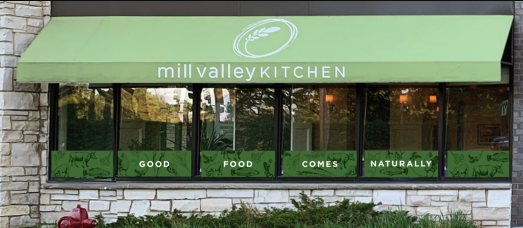 Green awning with mill valley KITCHEN above restaurant windows at 3805 Inglewood Ave S St Louis Park MN. Windows display GOOD FOOD COMES NATURALLY, with green panels of animal and plant art, plus stone wall and green plants in front.