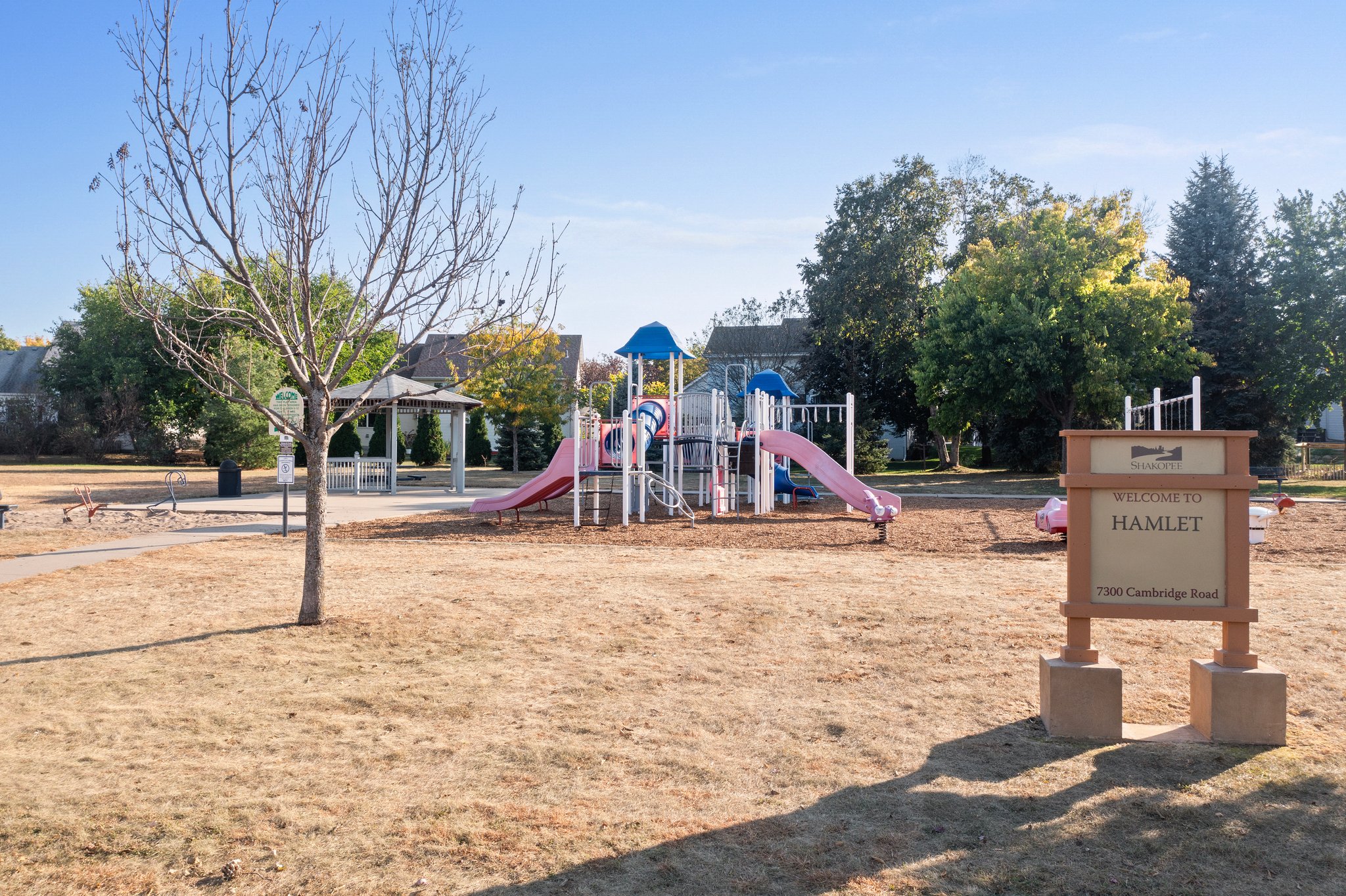 A playground with pink and blue slides and climbing structures sits on dry grass, surrounded by trees near Whitehall Place. A sign in the foreground reads “Welcome to Hamlet, 7307 Cambridge Road.” The clear blue sky completes the scene.