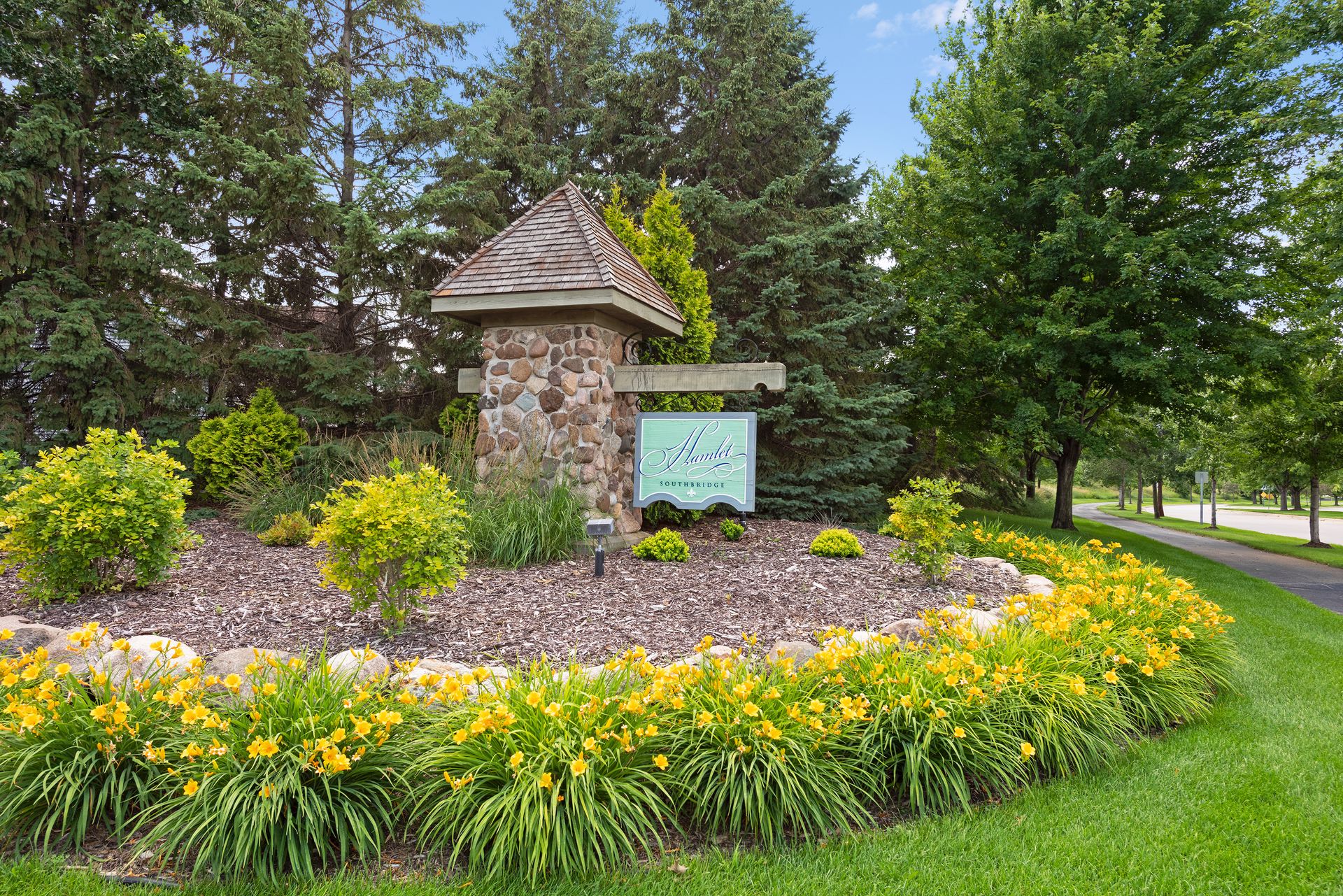A landscaped entrance features a stone pillar with a wooden roof and a sign reading Hawks Landing. Yellow flowers, green shrubs, and trees surround the area, as seen in Whitehall Place photos, with a road visible to the right.