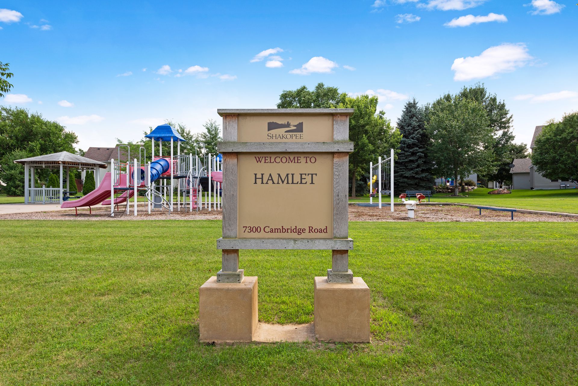 A welcome sign for Hamlet neighborhood at 7300 Cambridge Road stands on grass in front of a playground, with slides and climbing structures, while trees and a blue sky complete the scene—reminiscent of Whitehall Place in community PHOTOS.