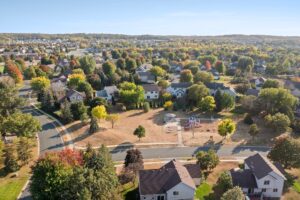 Aerial view of Whitehall Place suburban neighborhood with houses, tree-lined streets, and a small park featuring a playground and open grassy area. Trees show a mix of green and autumn colors under a clear blue sky. PHOTOS,1648.