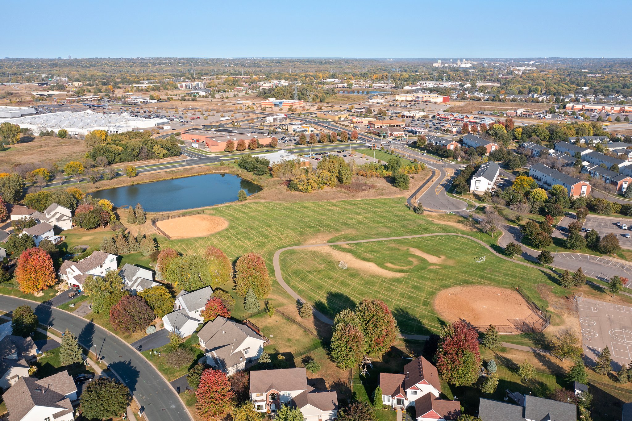 Aerial view PHOTOS of Whitehall Place, a suburban neighborhood with houses, two baseball fields, a pond, parking lot, and commercial buildings in the background, surrounded by trees and roads on a clear day. Image ID: 1648.