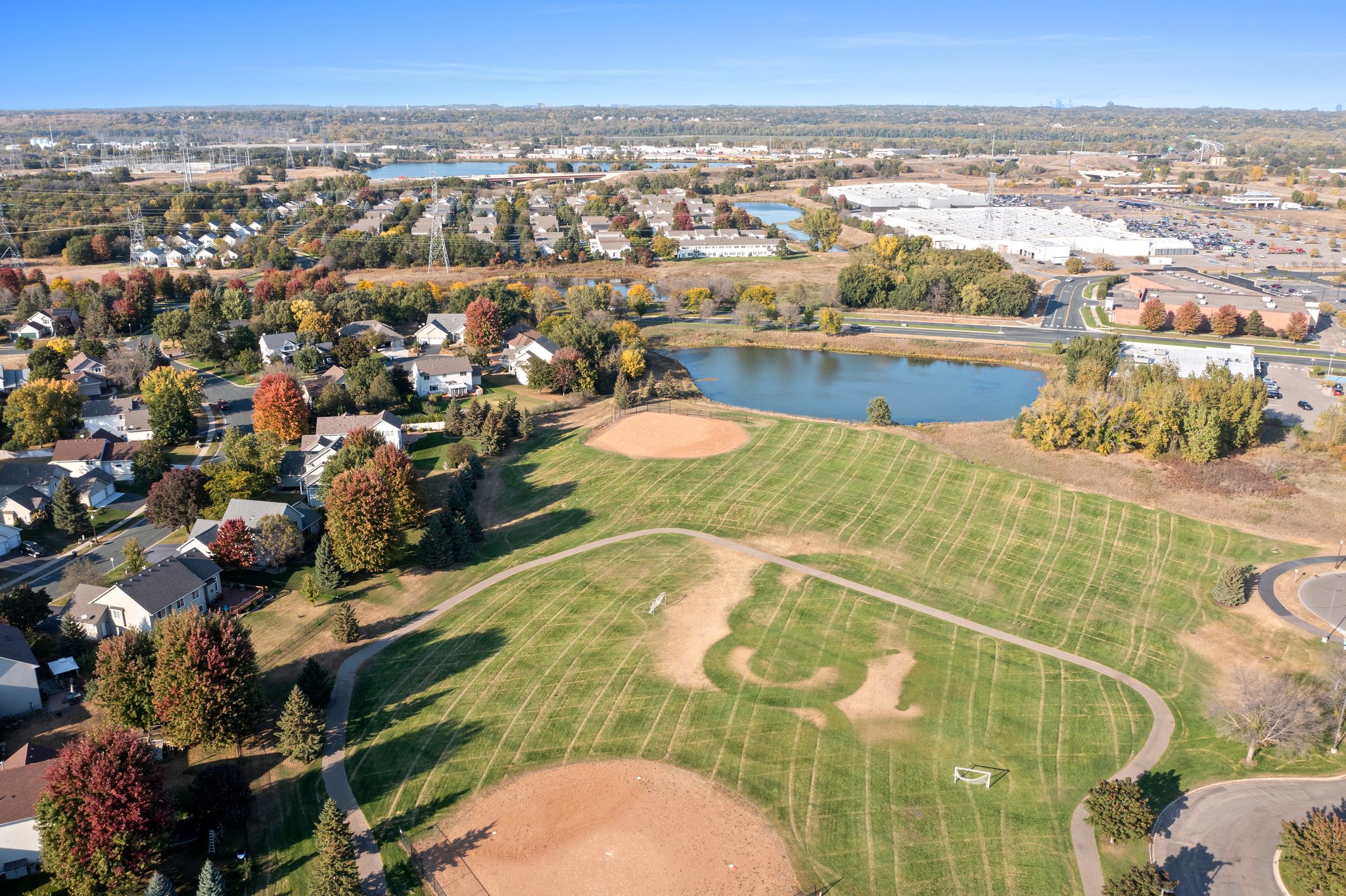 Aerial view of Whitehall Place neighborhood with baseball fields, a pond, autumn foliage trees, houses, and a nearby shopping center in the background under a clear sky. Stunning photos capture this vibrant suburban scene.