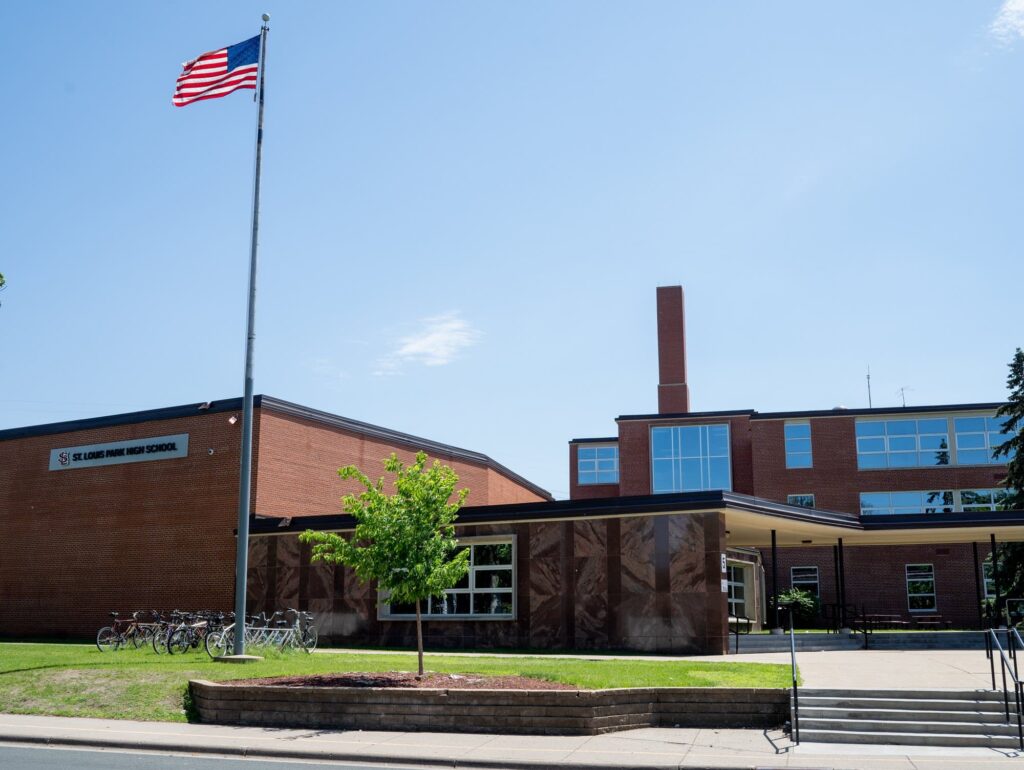 A brick high school building with large windows, an American flag on a tall pole, and several bicycles parked outside on a sunny day at 3805 Inglewood Ave S, St Louis Park, MN. The sign reads St. Cloud Area High School.