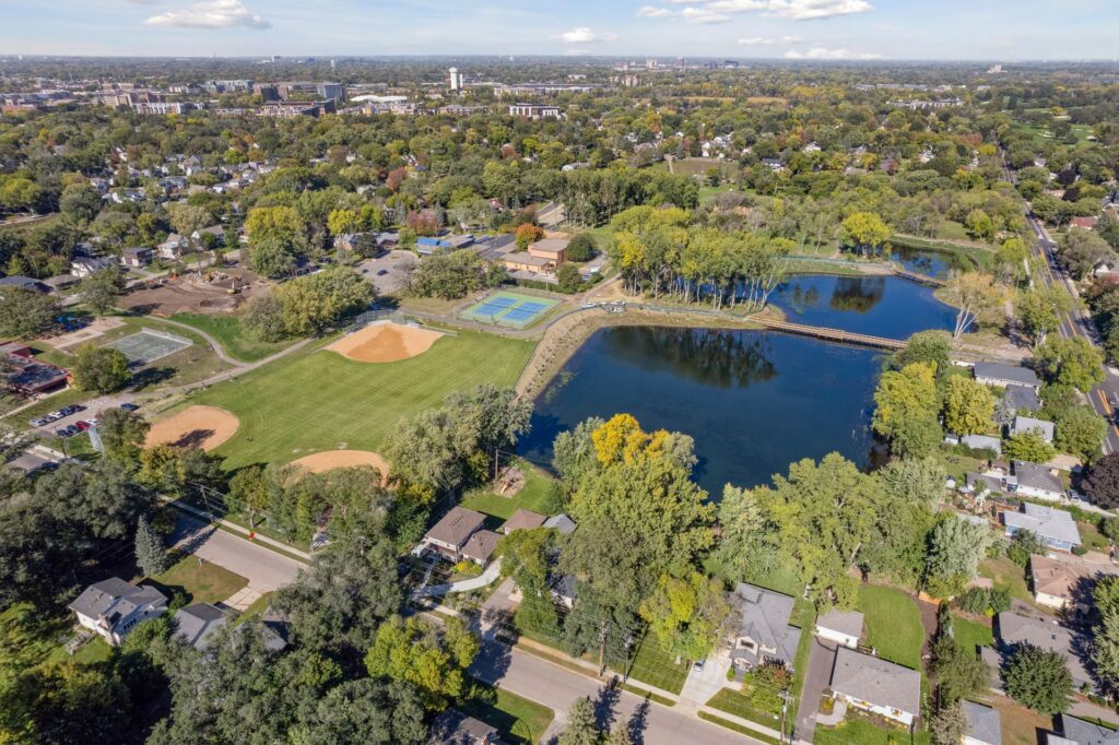 Aerial view of a suburban neighborhood with a baseball field, tennis courts, a large pond, and surrounding trees, with houses and distant city buildings in the background under a blue sky.