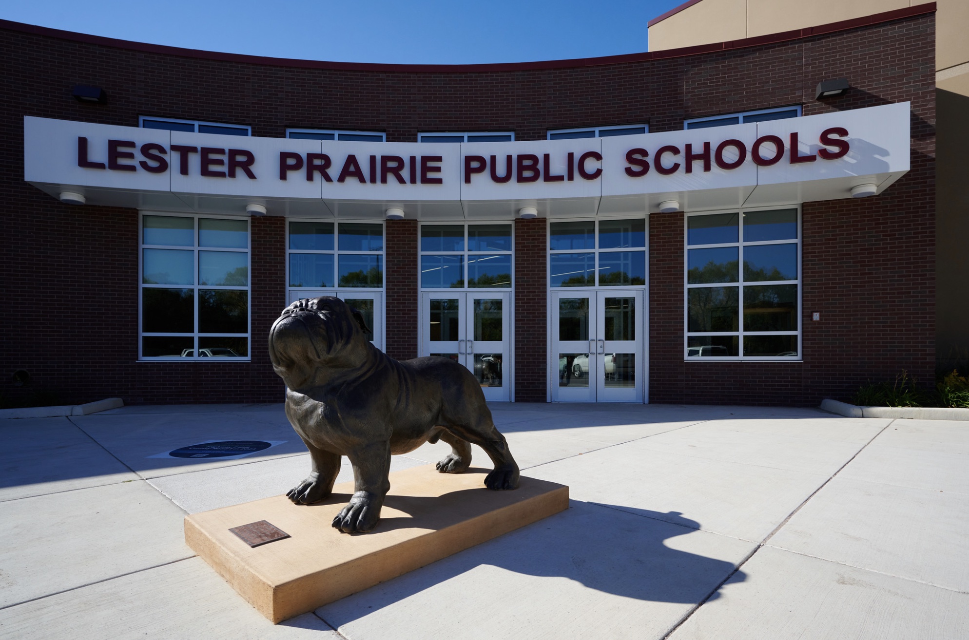 A bronze bulldog statue stands on a pedestal in front of the glass entrance to Lester Prairie Public Schools at 236 Pine St, Lester Prairie, MN, with large lettering above the doors and a brick building facade in the background.