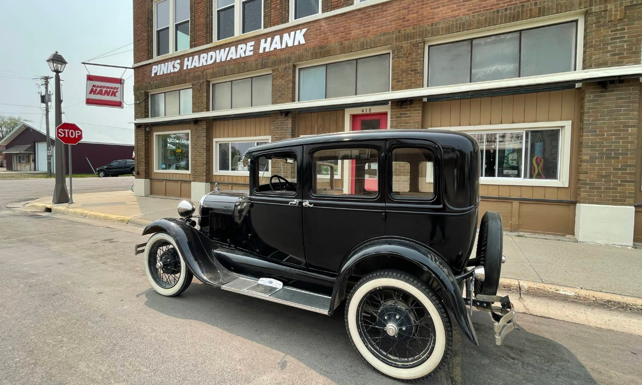 A black vintage car with whitewall tires is parked on a street in front of a brick building at 236 Pine St Lester Prairie MN, with PINKS HARDWARE HANK on the sign above the entrance and a stop sign on the corner.