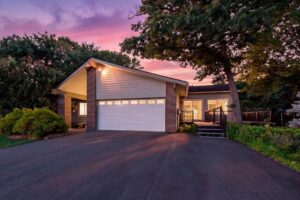 A modern house with a two-car garage, brick and siding exterior, front porch, and surrounding greenery, pictured at sunset with a purple and pink sky.