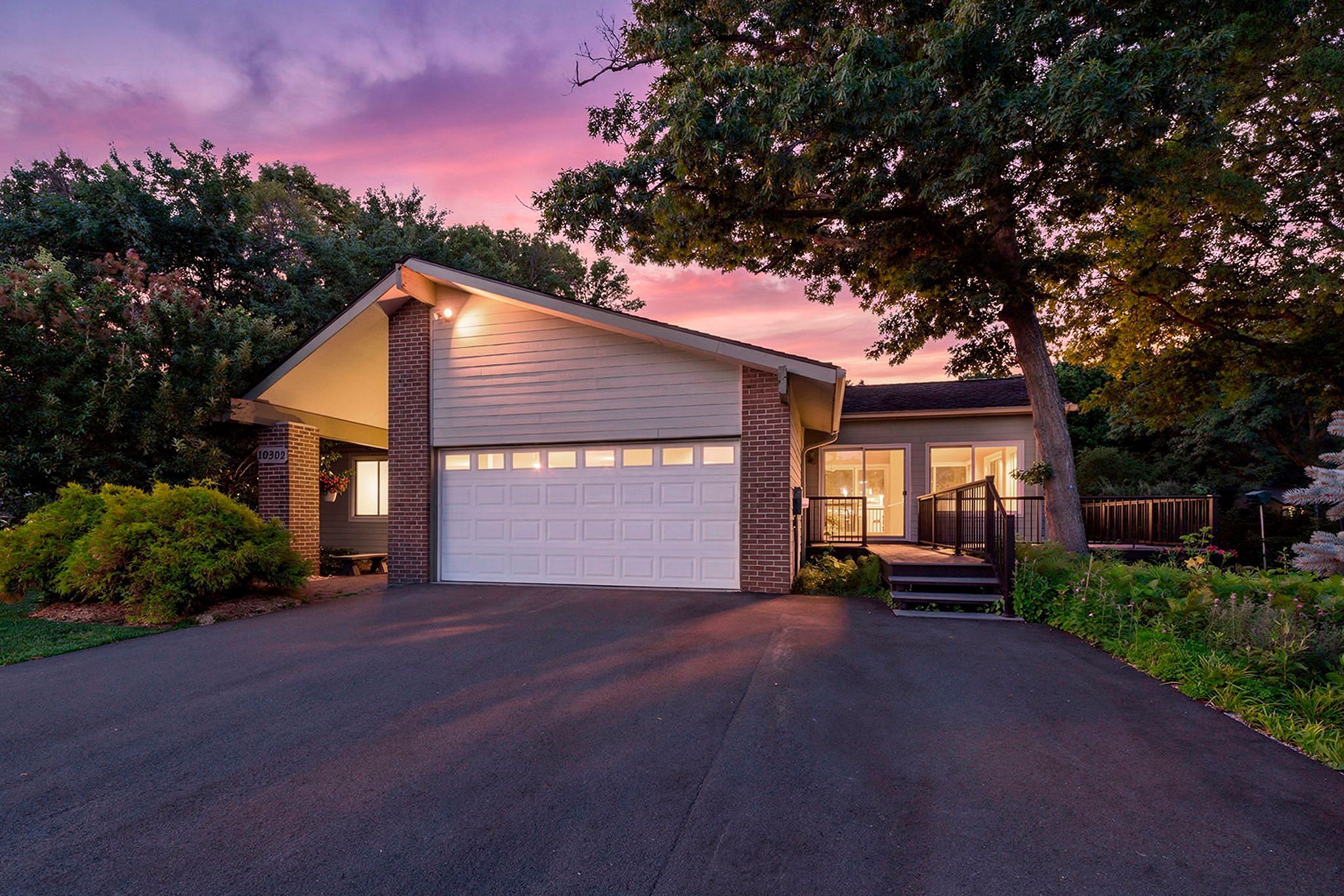 A modern house with a two-car garage, brick and siding exterior, front porch, and surrounding greenery, pictured at sunset with a purple and pink sky.