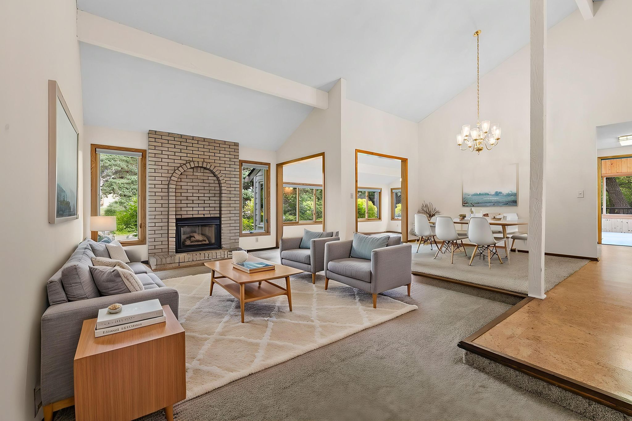 A modern living and dining area with neutral decor, large windows, a brick fireplace, gray sofas, a wooden coffee table, and a dining table with white chairs under a chandelier.