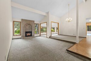 Spacious living room with gray carpet, brick fireplace, large windows, a raised dining area with chandelier, and views of greenery outside, connecting to the kitchen.