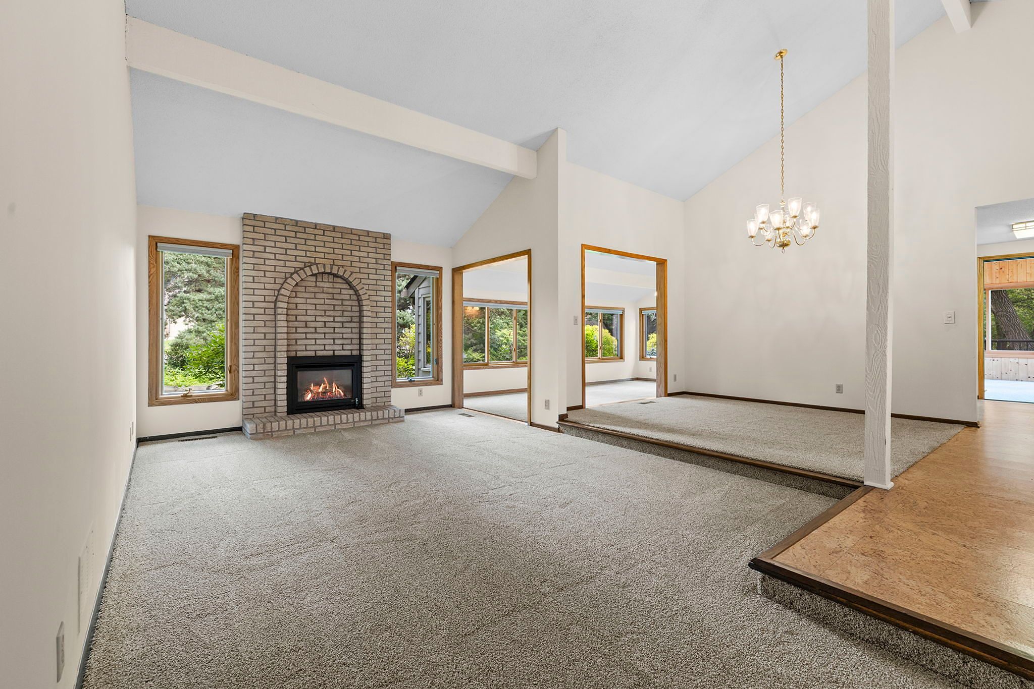 Spacious living room with gray carpet, brick fireplace, large windows, a raised dining area with chandelier, and views of greenery outside, connecting to the kitchen.