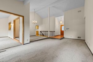 Spacious living room with vaulted ceiling, light carpet, white walls, two chandeliers, and a view of the entryway, staircase, and adjacent room with wood trim and carpet.