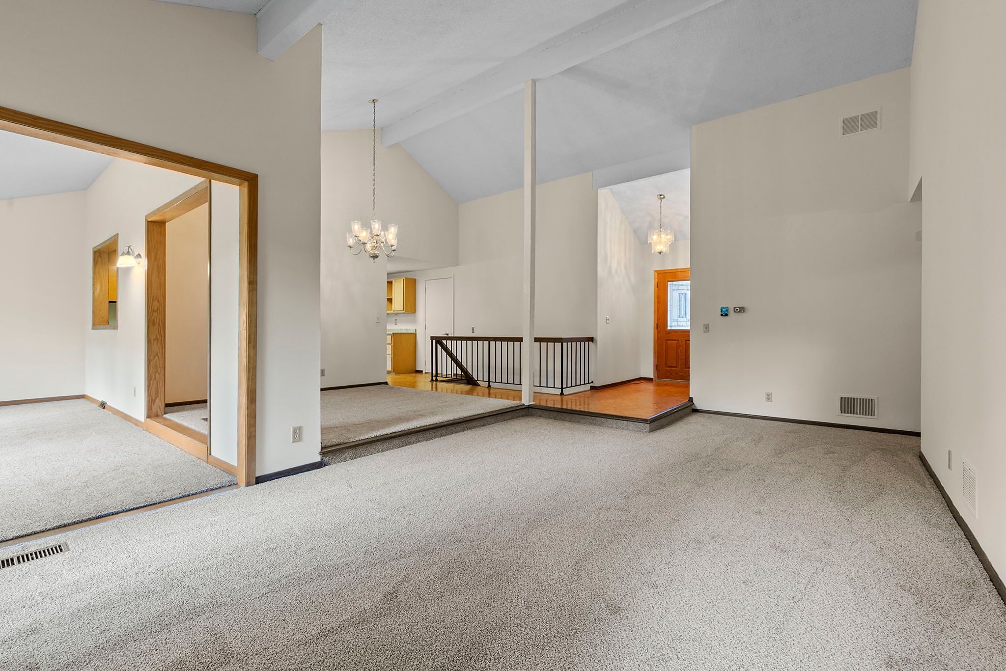 Spacious living room with vaulted ceiling, light carpet, white walls, two chandeliers, and a view of the entryway, staircase, and adjacent room with wood trim and carpet.