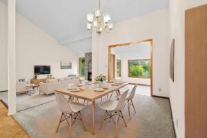 A modern dining room with a wooden table set for four, white chairs, a chandelier above, and an open view into a bright living room and sunroom with large windows showing greenery outside.