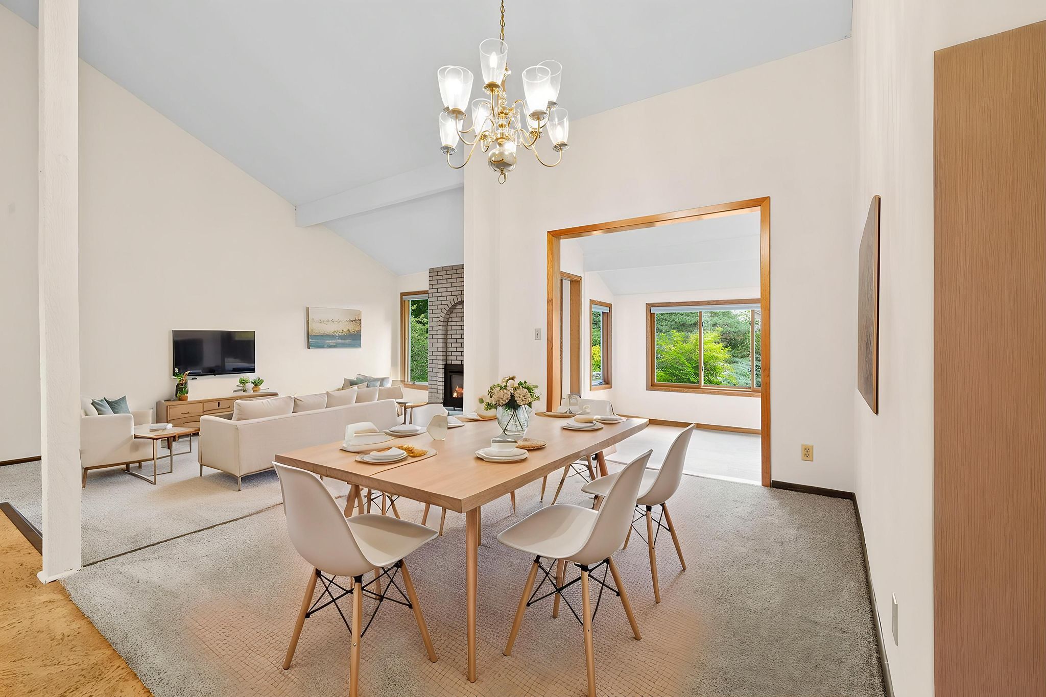 A modern dining room with a wooden table set for four, white chairs, a chandelier above, and an open view into a bright living room and sunroom with large windows showing greenery outside.
