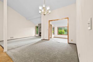 Spacious, carpeted living room with chandelier, a view into a sunroom with large windows, light walls, and a brick fireplace. Natural light fills both rooms.