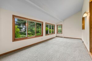 A long, empty room with beige carpet, white walls, large windows with wooden trim, and a sloped ceiling. Green trees and bushes are visible outside the windows.