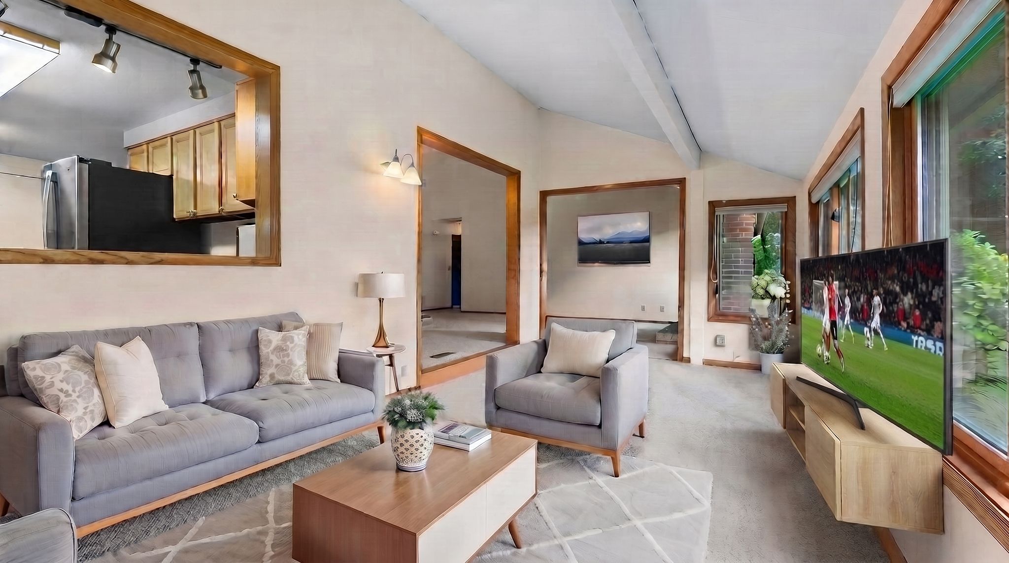Modern living room with gray sofa, armchair, coffee table, TV showing a soccer game, large windows, beige walls, open doorway to kitchen, and a light carpet. Natural light fills the space.