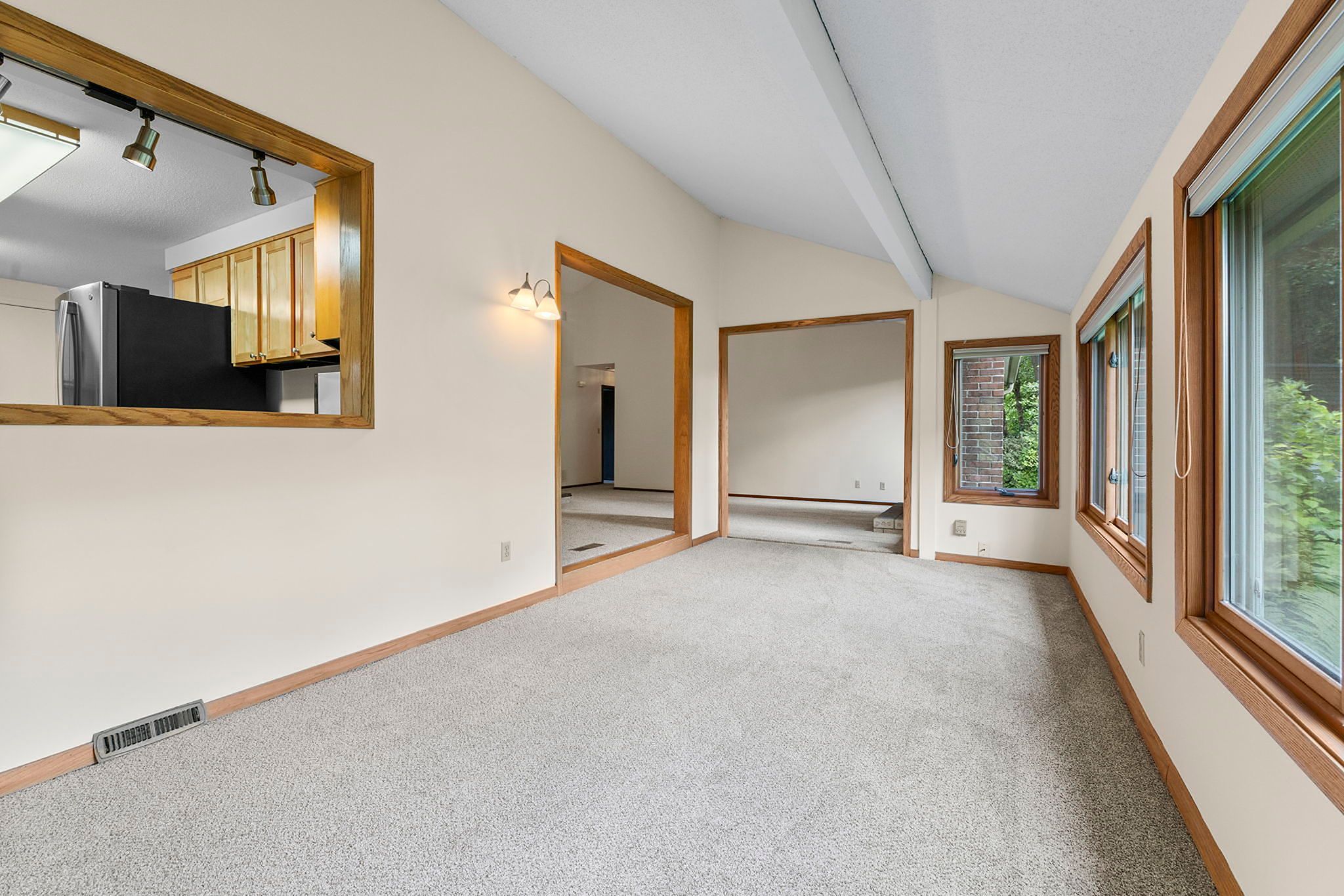 Spacious carpeted room with cream walls, large windows on the right, and a view into the kitchen through a wood-framed opening. Recessed lighting and natural light brighten the space.