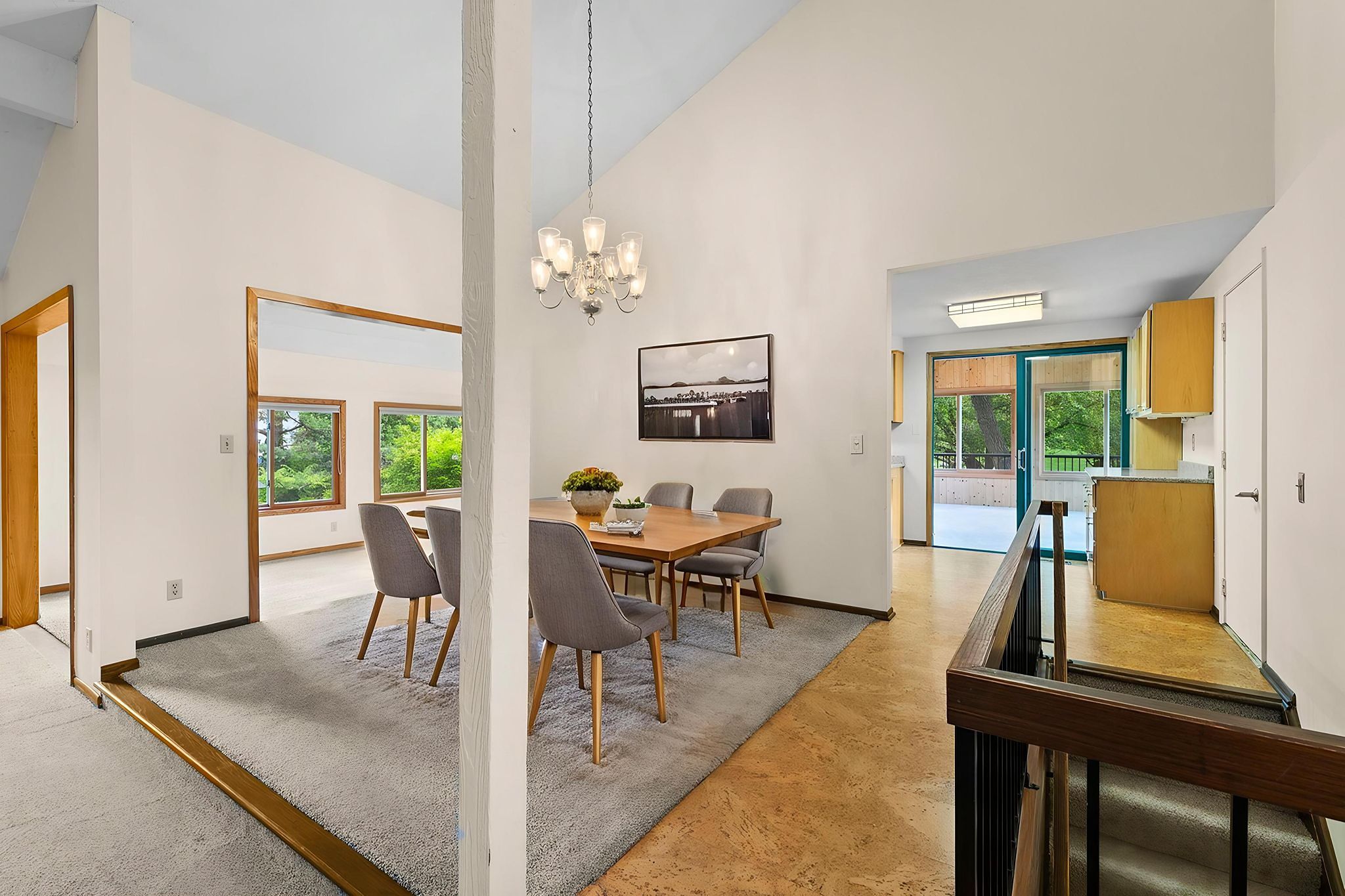Dining area with a wooden table and gray chairs on a rug, modern chandelier above, and open view into a kitchen and living room with large windows and natural light.