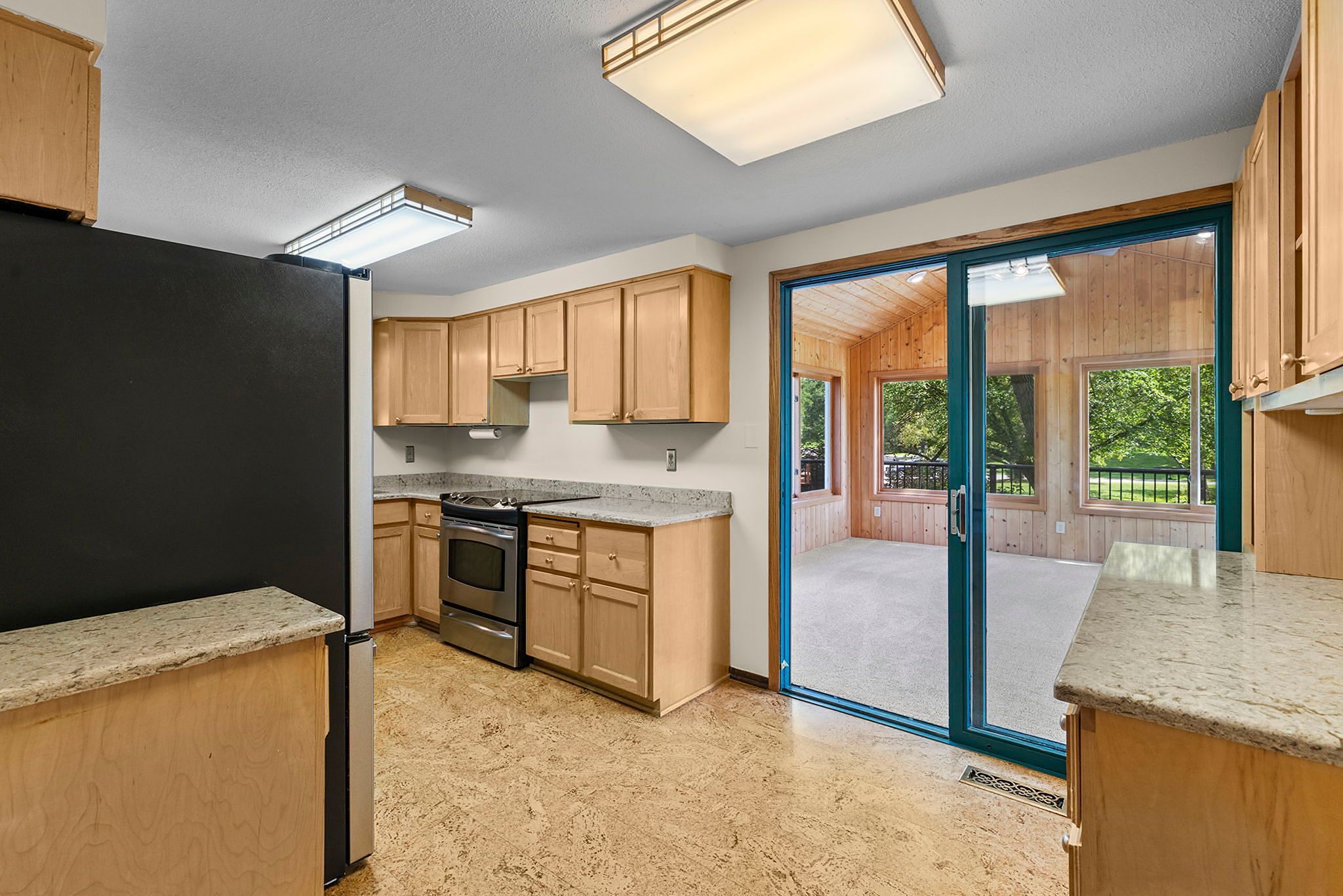 Modern kitchen with light wood cabinets, granite countertops, stainless steel appliances, and sliding glass doors leading to a sunroom with large windows showing green trees outside.