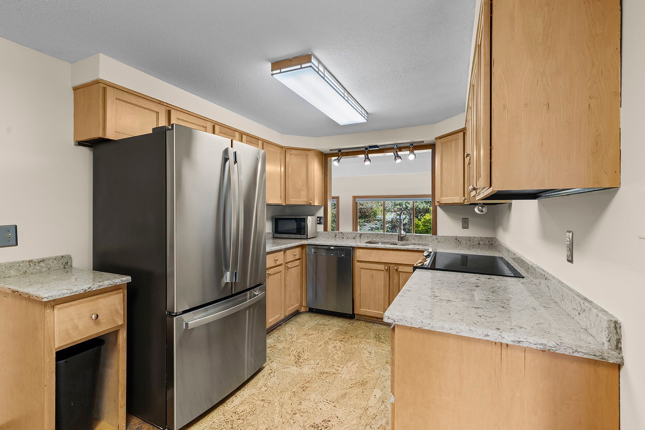 Modern kitchen with light wood cabinets, stainless steel fridge, dishwasher, and microwave, granite countertops, electric stove, large window over the sink, and overhead lighting.