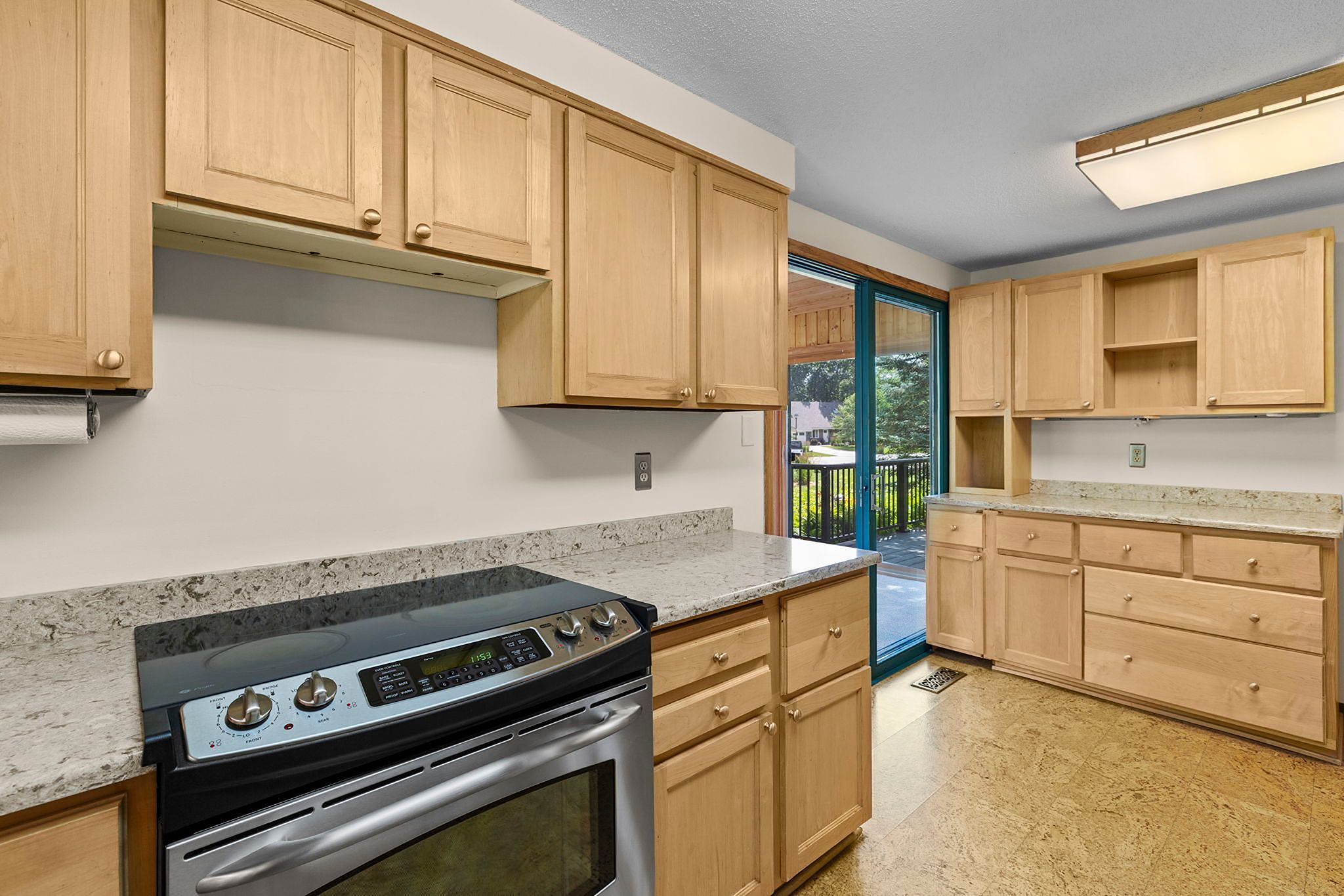 Modern kitchen with light wood cabinets, stainless steel oven, marble countertops, and large sliding glass doors leading to an outdoor deck. Natural light brightens the space.