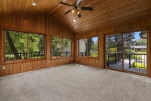 A sunroom with wood-paneled walls and ceiling, carpeted floor, large windows, and a ceiling fan. Sliding glass doors open to a deck with a view of trees and neighboring houses.