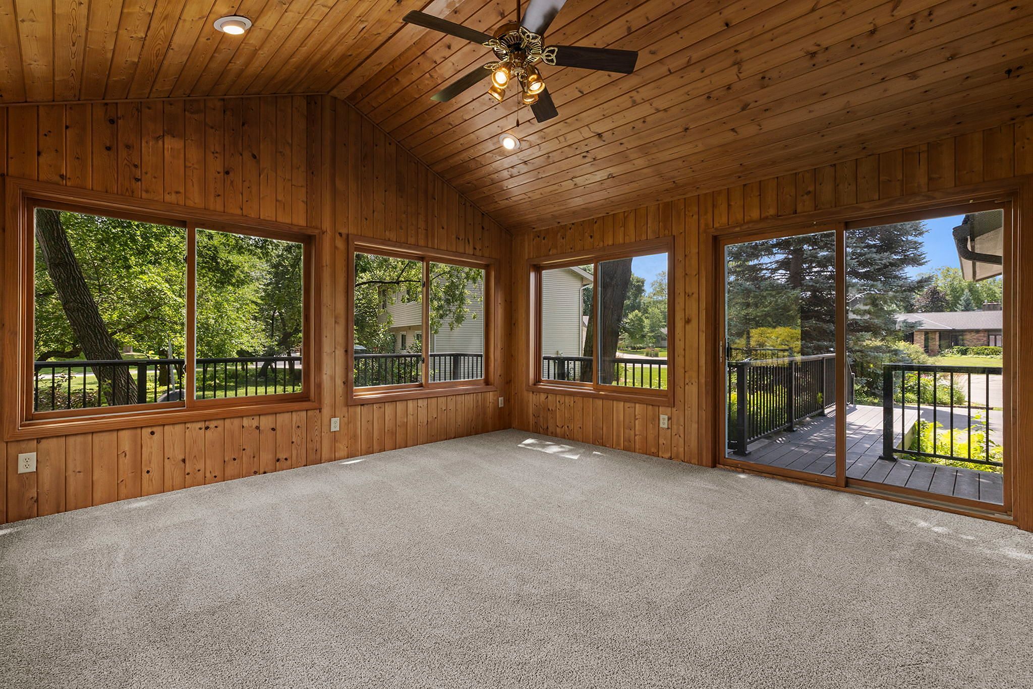 A sunroom with wood-paneled walls and ceiling, carpeted floor, large windows, and a ceiling fan. Sliding glass doors open to a deck with a view of trees and neighboring houses.