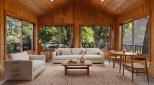 A cozy sunroom with wood-paneled walls and ceiling, large windows, beige sofa, armchair, wooden coffee table, dining set, and a potted plant. Natural light fills the space and greenery is visible outside.