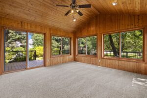 A sunroom with wooden walls and ceiling, large windows overlooking a green yard, beige carpet, a ceiling fan with lights, and a door leading to an outdoor deck.