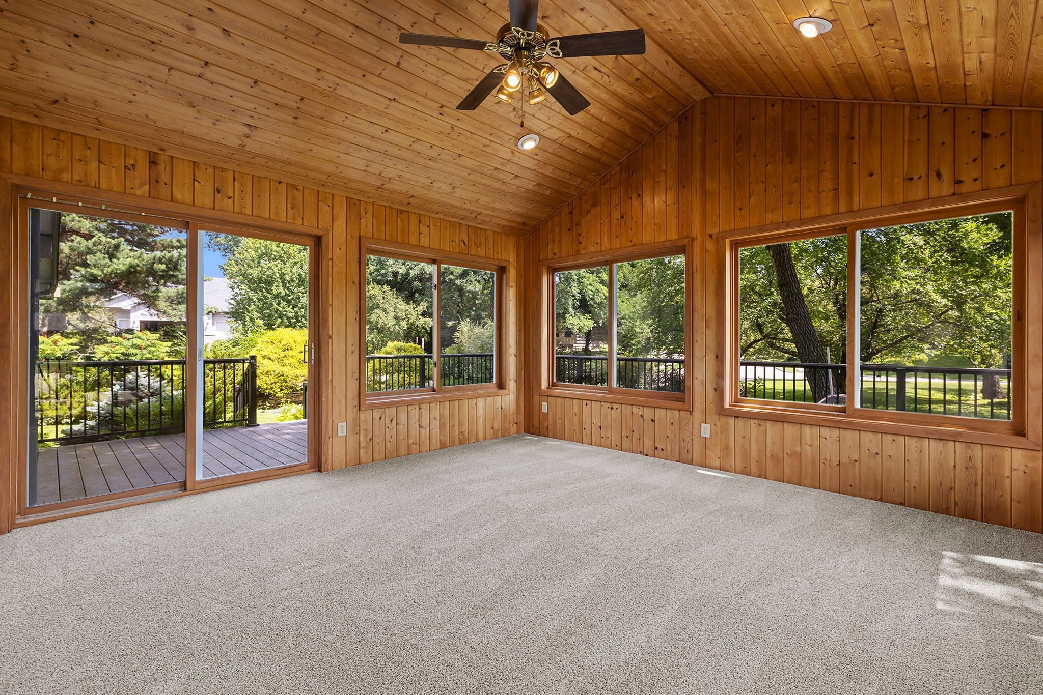 A sunroom with wooden walls and ceiling, large windows overlooking a green yard, beige carpet, a ceiling fan with lights, and a door leading to an outdoor deck.