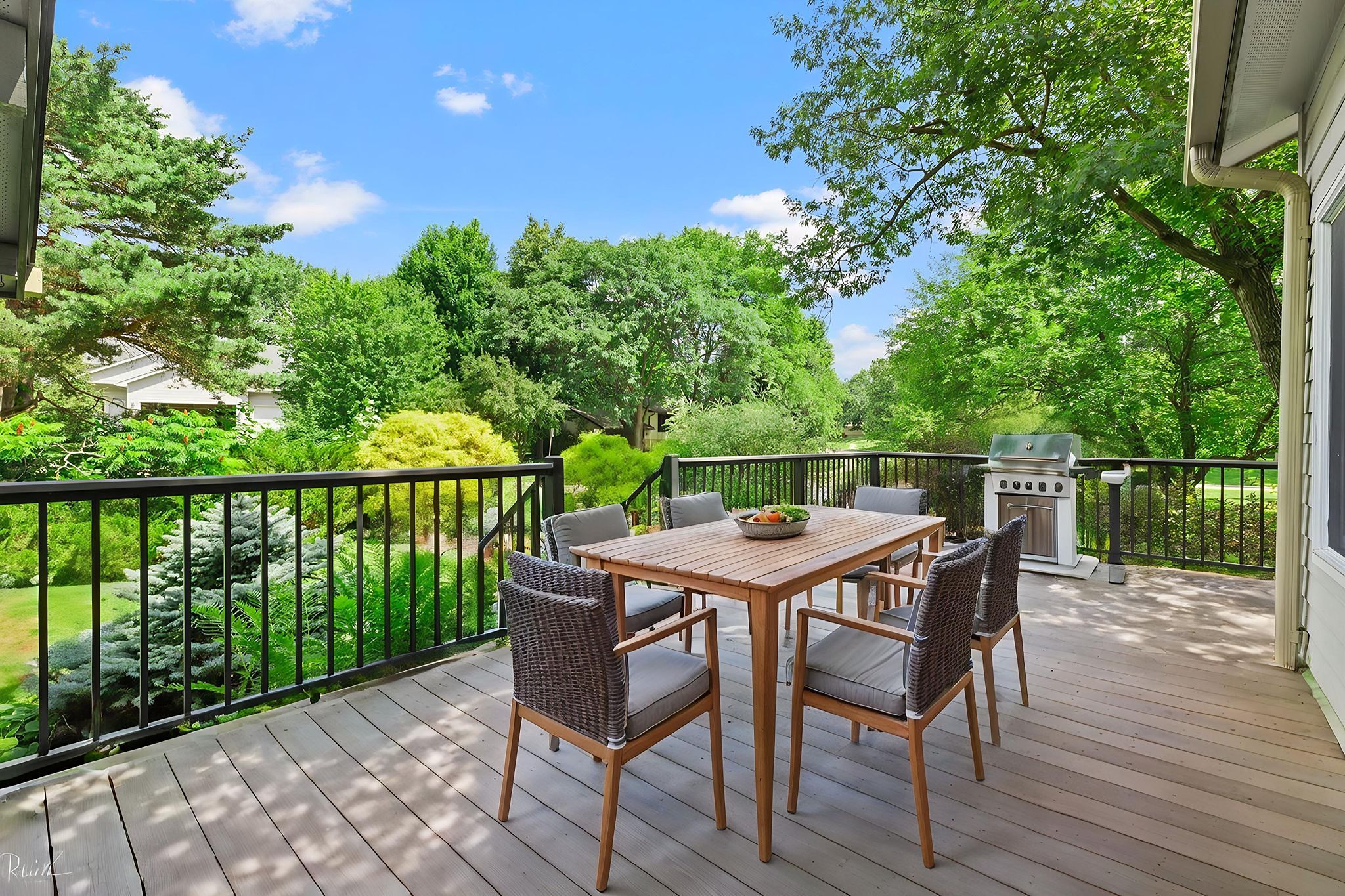 A wooden deck with a table, six chairs, and a grill overlooks a lush, green backyard with trees and shrubs under a bright blue sky.