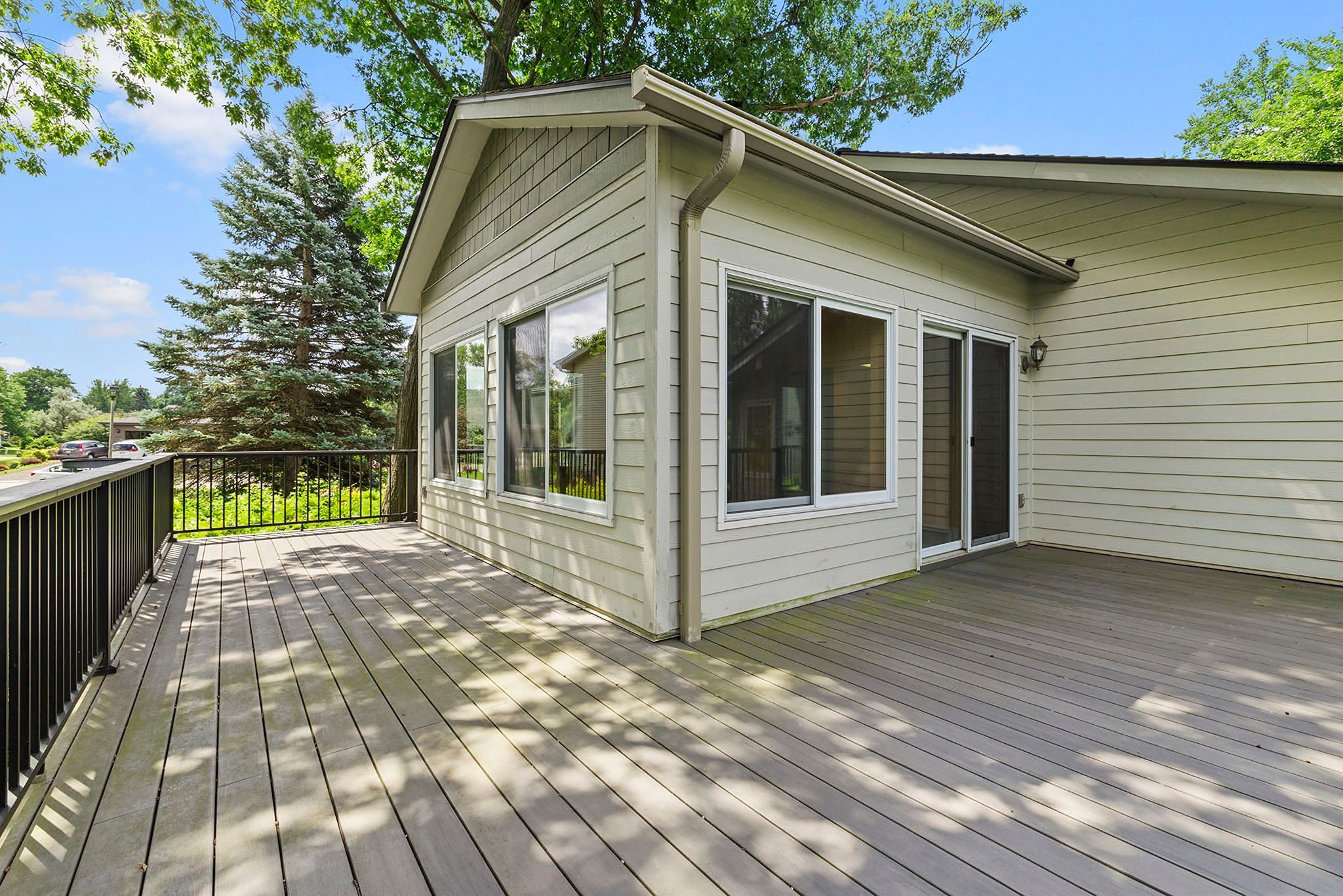 Spacious wooden deck outside a beige house, featuring large windows and black metal railing, surrounded by trees and greenery under a bright blue sky.