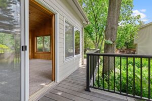 A wooden deck with black railings outside a house; a sliding glass door opens to a carpeted room with wood-paneled walls. Lush green trees and a neighboring house are visible in the background.