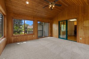A sunroom with wood-paneled walls and ceiling, large windows, a ceiling fan with lights, carpeted floor, and a sliding glass door overlooking a patio and greenery outside.